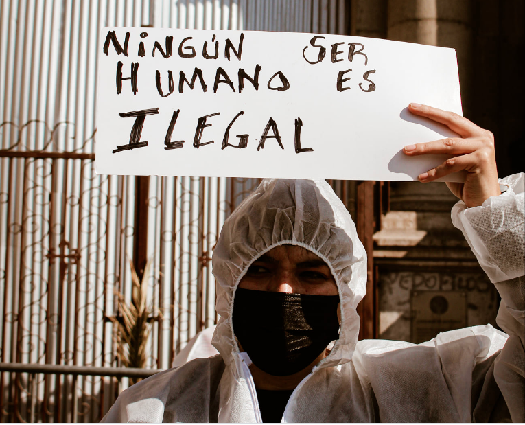 Persona en overol blanco con mascarilla sostiene un letrero "ningún ser humano es ilegal"