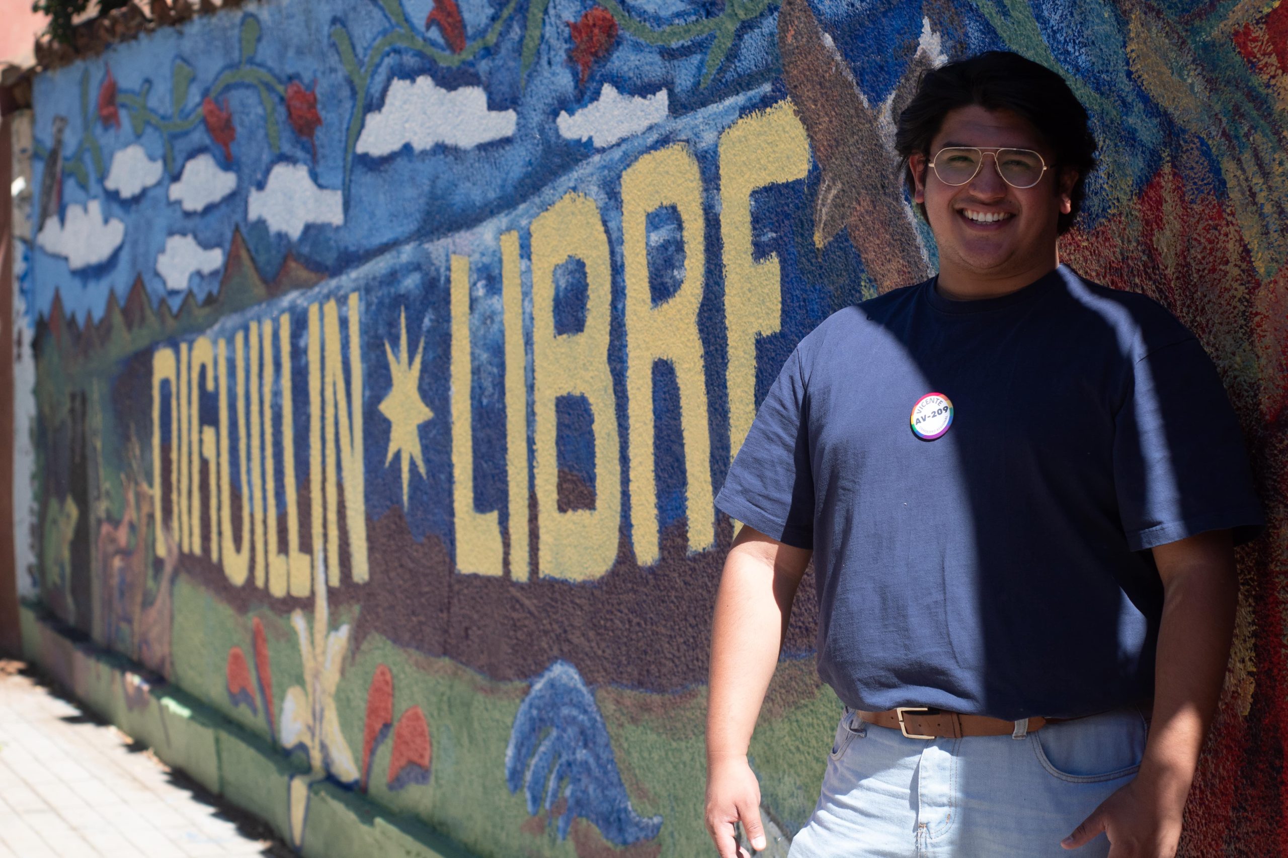 Un joven sonriente está de pie ocupando el lado derecho de la pantalla, está apoyado en un mural que dice "Diguillín libre"
