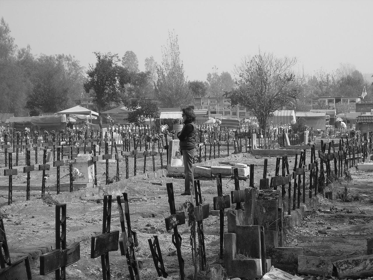 Imagen de una persona parada en un cementerio: Hay varias cruces de aspecto descuidado marcando el lugar de las tumbas