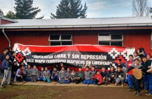 Decenas de niños y voluntarios de la Red por la Defensa de la Infancia Mapuche posan frente a un lienzo rojo y negro que exige "Infancia libre y sin represión".